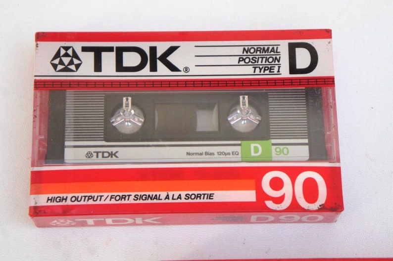 Puede incluir: Una caja de cassette de audio roja, blanca y negra con el texto "TDK" y "D 90" impreso en ella. La caja est&aacute; etiquetada "Posici&oacute;n Normal Tipo I" y "Alta Salida/Se&ntilde;al Fuerte a la Salida".
