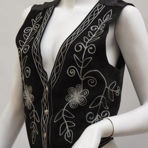 May include: Black vest with silver floral embroidery. The vest features a V-neck, button closure, and decorative silver stitching along the edges and around the floral designs. The vest is displayed on a mannequin.