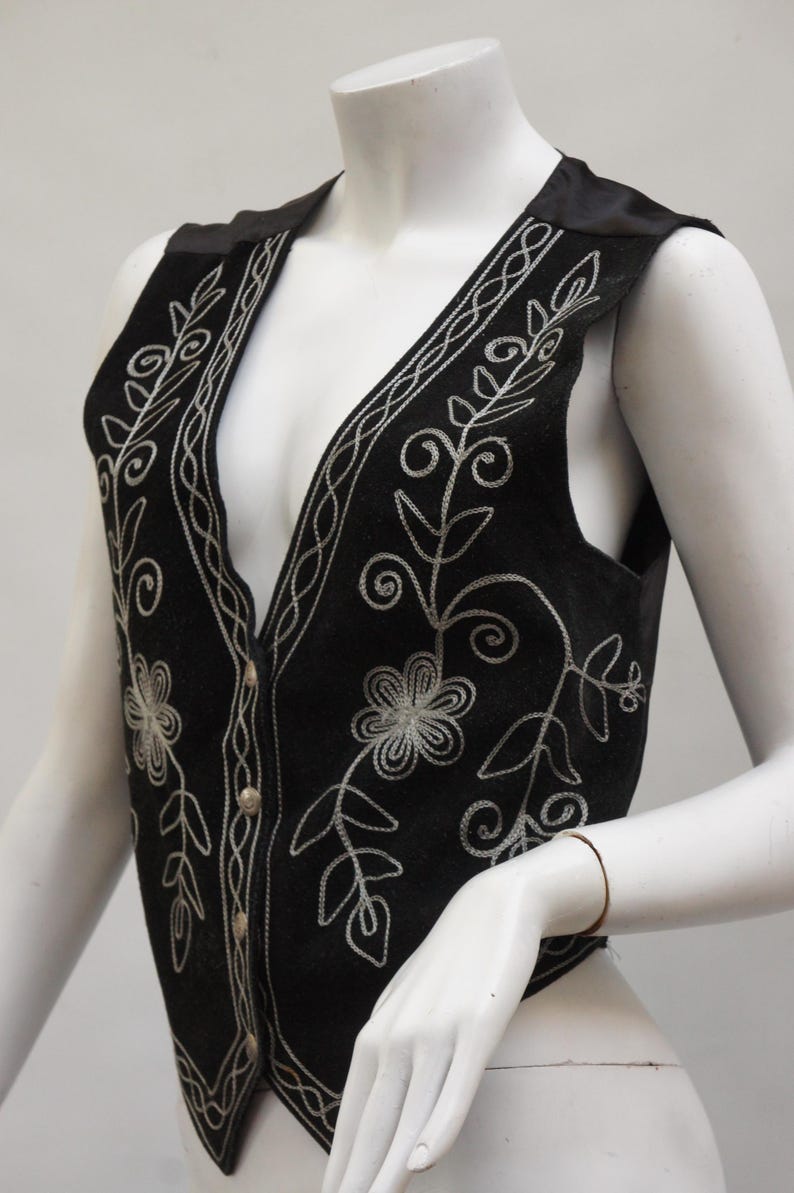 May include: Black vest with silver floral embroidery. The vest features a V-neck, button closure, and decorative silver stitching along the edges and around the floral designs. The vest is displayed on a mannequin.