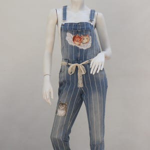 Op de afbeelding: Blauwe denim overall met witte strepen en geborduurde kattenontwerpen. De overall heeft een witte kantenriem en een ontspannen pasvorm.