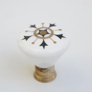 Könnte beinhalten: Weißer Keramik-Schrankknopf mit dekorativem schwarz-goldenen Design. Der Knopf hat einen Messingfuß und eine abgerundete Oberseite mit einem blütenartigen Muster. Das Design ist auf der Oberseite des Knopfes zentriert.