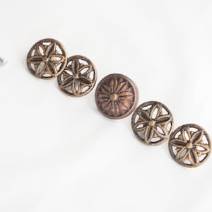 Könnte beinhalten: Vier runde Schubladenknöpfe aus bronzefarbenem Metall mit durchbrochenem Blumenmuster und ein massiver mittlerer Knopf mit strukturiertem Blütenblattdesign, begleitet von vier silberfarbenen Metallschrauben zur Montage.