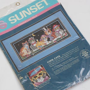 May include: A Sunset counted cross stitch kit featuring a design of "Cool Cats" wearing hats and sunglasses. The kit includes a color-accented chart and is packaged in a clear plastic sleeve. The finished size is 10" x 11".