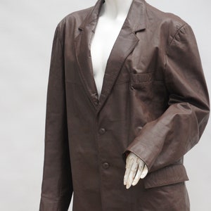 May include: A brown leather blazer with a notched lapel, two-button closure, and a chest pocket. The jacket is displayed on a mannequin. The blazer has a classic, tailored design.