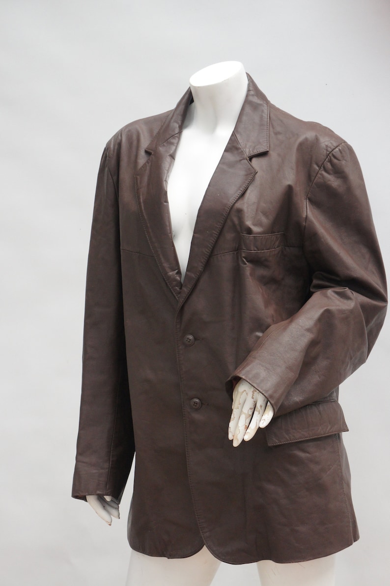 May include: A brown leather blazer with a notched lapel, two-button closure, and a chest pocket. The jacket is displayed on a mannequin. The blazer has a classic, tailored design.