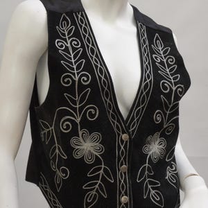 May include: Black vest with silver floral embroidery. The vest features a V-neck and a button-down front. The floral design includes flowers and leaves, with a decorative border along the edges.