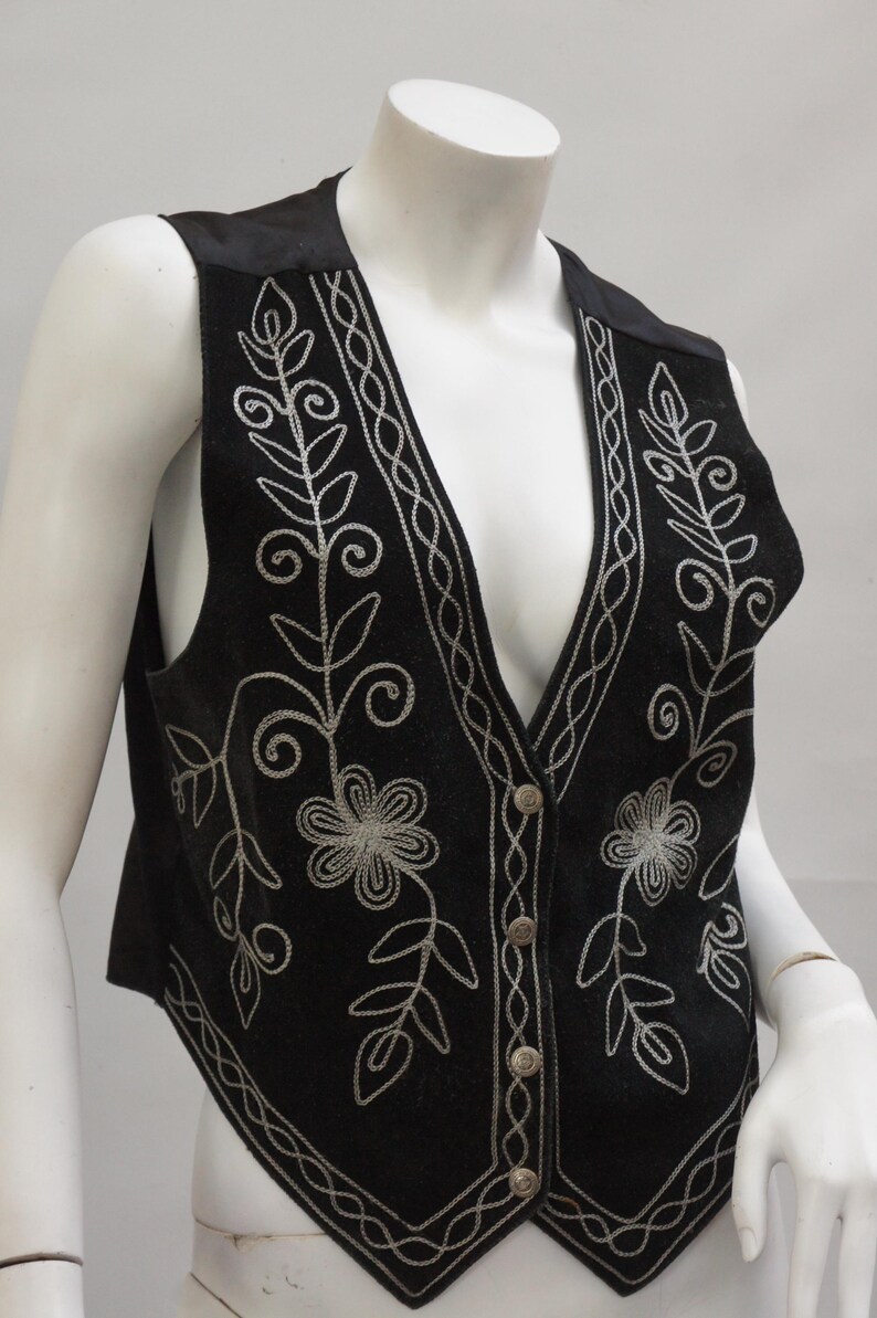 May include: Black vest with silver floral embroidery. The vest features a V-neck and a button-down front. The floral design includes flowers and leaves, with a decorative border along the edges.