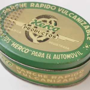 May include: A green and yellow oval tin with the text "PARCHE RAPIDO VULCANIZABLE" and "PRODUCTOS "HERCO" PARA EL AUTOMOVIL" printed on it. The tin has a small, round, metal lid with a series of small, round holes.