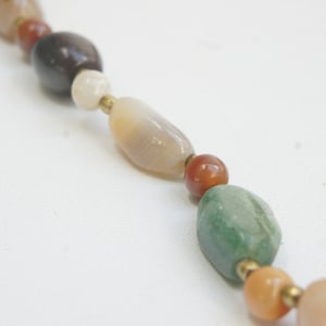 May include: A close-up of a multi-colored gemstone necklace. The necklace features oval-shaped beads in shades of brown, white, green, and orange.