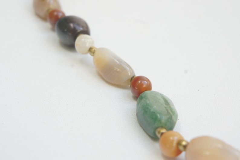 May include: A close-up of a multi-colored gemstone necklace. The necklace features oval-shaped beads in shades of brown, white, green, and orange.