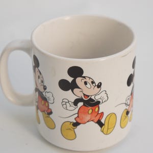 以下が含まれることがあります： ミッキーマウスのデザインが施された白いセラミックマグカップ。マグカップには、赤いショートパンツと黄色の靴を履いたミッキーマウスが歩いている様子が4つ描かれています。