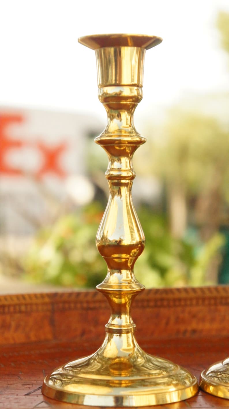May include: A pair of gold metal candle holders with a classic design. The holders have a round base and a tall, slender stem with a decorative, fluted design.