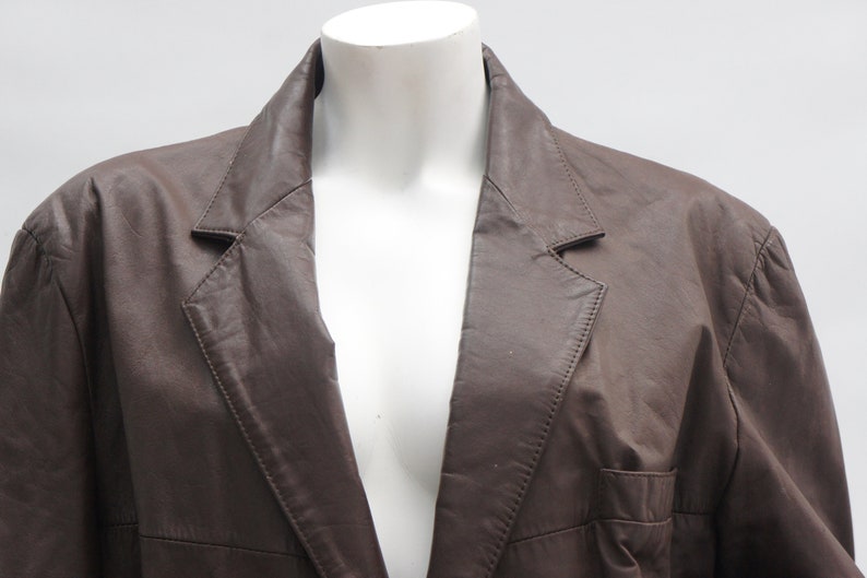 May include: A brown leather blazer with a notched lapel and a single-breasted design. The jacket has a classic tailored fit and a pocket on the front. The blazer is displayed on a white mannequin.