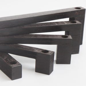 May include: A set of dark brown wooden candle holders arranged in a staggered formation. Each holder has a rectangular base and a horizontal top piece with a circular hole for a candle. The wood grain is visible.