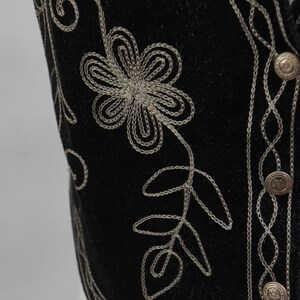 May include: Close-up of a black vest with intricate silver floral embroidery. The vest features a detailed flower and leaf design, along with decorative silver stitching along the edges and around the buttons. The vest has three silver buttons.