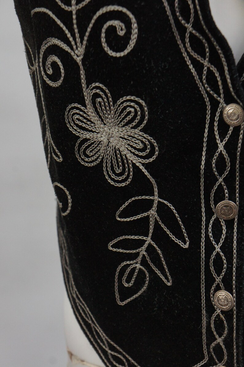 May include: Close-up of a black vest with intricate silver floral embroidery. The vest features a detailed flower and leaf design, along with decorative silver stitching along the edges and around the buttons. The vest has three silver buttons.