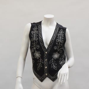 May include: Black vest with silver floral embroidery. The vest has a V-neck and a button-up front. The embroidery features floral and leaf designs. The vest is displayed on a mannequin.