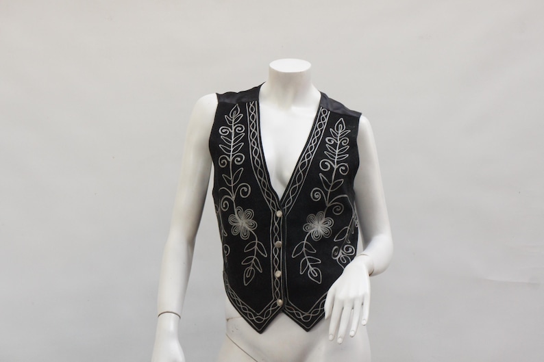 May include: Black vest with silver floral embroidery. The vest has a V-neck and a button-up front. The embroidery features floral and leaf designs. The vest is displayed on a mannequin.
