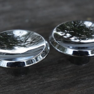Op de afbeelding: Twee glanzende, verchroomde kastknoppen. Elke knop heeft een afgeronde, licht concave bovenkant met een reflecterend oppervlak en een korte, cilindrische basis. De knoppen staan tegen een donkere, getextureerde achtergrond.