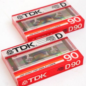 Puede incluir: Dos cintas de cassette de audio TDK rojas y blancas. Ambas cintas tienen una duraci&oacute;n de 90 minutos y est&aacute;n etiquetadas como "D 90". La cinta superior tiene el texto "HIGH OUTPUT/FORT SIGNAL A LA SORTIE" impreso en ella. La cinta inferior tiene el texto "NORMAL POSITION TYPE I" impreso en ella.
