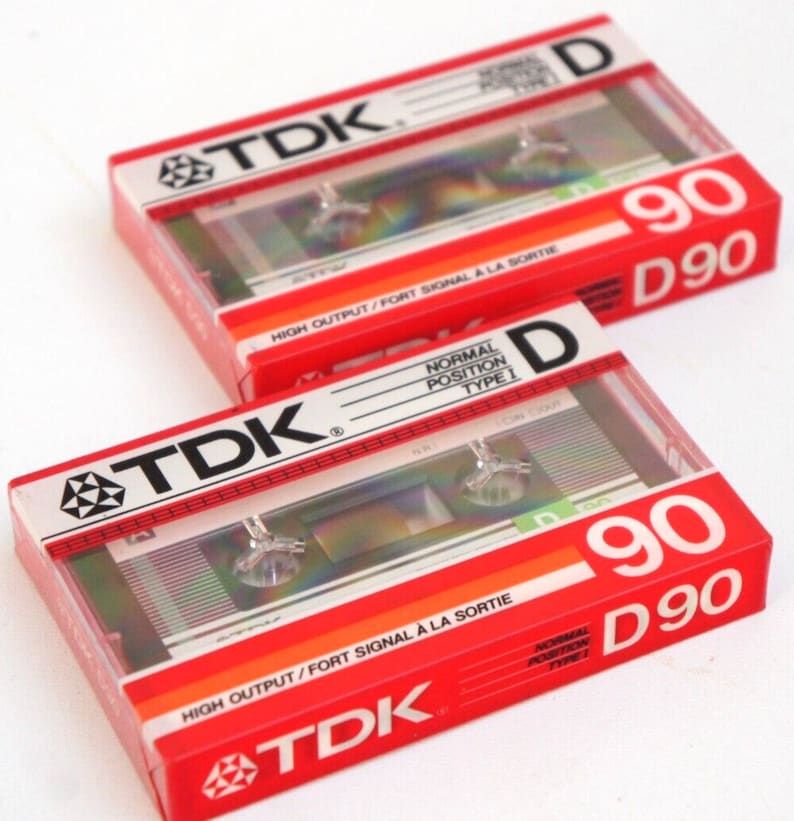 Puede incluir: Dos cintas de cassette de audio TDK rojas y blancas. Ambas cintas tienen una duraci&oacute;n de 90 minutos y est&aacute;n etiquetadas como "D 90". La cinta superior tiene el texto "HIGH OUTPUT/FORT SIGNAL A LA SORTIE" impreso en ella. La cinta inferior tiene el texto "NORMAL POSITION TYPE I" impreso en ella.