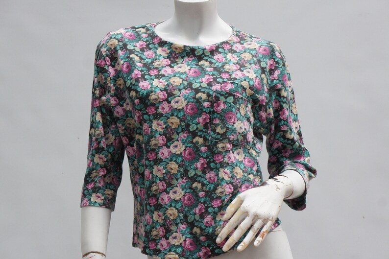 Puede incluir: Una blusa de estampado floral con fondo oscuro y flores rosas, blancas y verdes. La blusa tiene un bolsillo en el lado izquierdo y mangas tres cuartos.