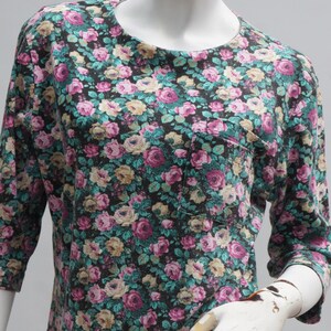 Puede incluir: Una camisa de estampado floral negro y turquesa con un peque&ntilde;o bolsillo en el lado izquierdo. La camisa tiene cuello redondo y mangas tres cuartos.