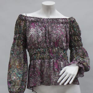 May include: Off-the-shoulder blouse with long sleeves. The sheer fabric features a floral pattern in shades of purple, green, and brown. The blouse has elastic at the neckline and waist, and is displayed on a mannequin.