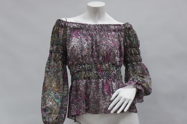 May include: Off-the-shoulder blouse with long sleeves. The sheer fabric features a floral pattern in shades of purple, green, and brown. The blouse has elastic at the neckline and waist, and is displayed on a mannequin.