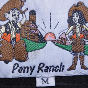 May include: Embroidered label with the text "Pony Ranch" and two cartoon cowboys. The cowboys are wearing cowboy hats, vests, and chaps. The label also features a sunrise and a size "M".