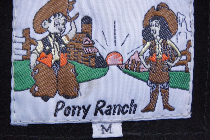 May include: Embroidered label with the text "Pony Ranch" and two cartoon cowboys. The cowboys are wearing cowboy hats, vests, and chaps. The label also features a sunrise and a size "M".