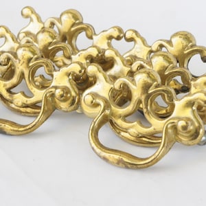 Op de afbeelding: Set van zes sierlijke, goudkleurige kastgrepen met een decoratief, rolvormig ontwerp. Elke greep heeft een eenvoudige, gebogen handgreep.