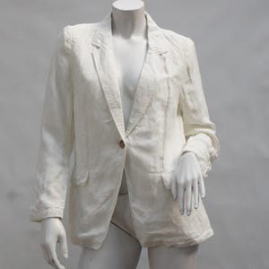 May include: A long-sleeved, off-white blazer featuring a notched lapel, a single front button closure, and two flap pockets at the waist. The garment is displayed on a mannequin, showcasing a structured collar and a straight hemline design.