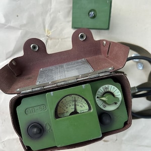 May include: A green DP-5B radiation survey meter housed in a brown leather case with a hinged lid. The device features a central analog gauge with Cyrillic text, a circular range selection dial, and a metal information plate on the inner lid.