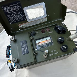 Peut inclure: Un radiamètre DP-12 vert olive avec un couvercle à charnière, une jauge analogique et des boutons de commande noirs. L'appareil présente des étiquettes en cyrillique, un petit miroir rectangulaire sur le couvercle intérieur et des fils électriques.