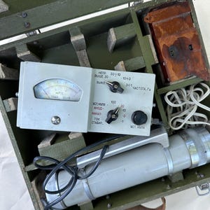 May include: A vintage Soviet-era radiation detection device housed in a green wooden carrying case. The unit features a light gray control panel with a needle gauge, two black rotary knobs, and Cyrillic text. A cylindrical probe is included.