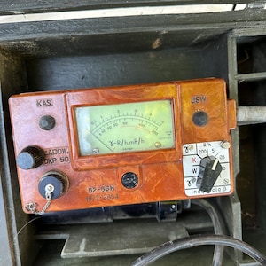 May include: A vintage DP-66M radiation dosimeter in a brown marbled plastic casing, featuring a central analog gauge with gamma R/h and mR/h scales, a black rotary selector switch, and text labels including KAS, USW, and DKP-50, housed in a dark green box.