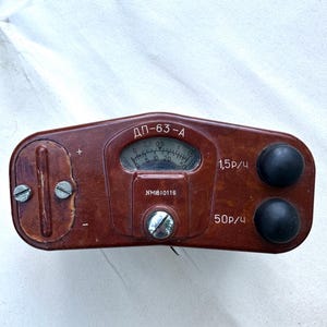 May include: A brown, rectangular radiation dosimeter device with rounded edges, featuring a central analog gauge, two black circular knobs on the right, and a vertical adjustment dial on the left. The text "ДП-63-А", "NMI610116", "1,5р/ч", and "50р/ч" are printed on the front surface.
