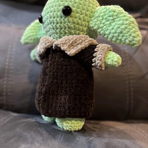 May include: A green crocheted plush toy with large black eyes, wearing a brown and gray crocheted coat. The toy is sitting on a gray couch.