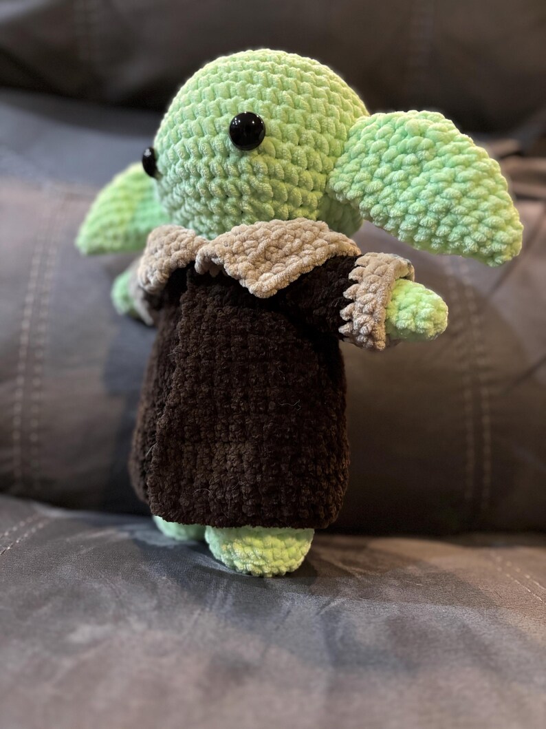 May include: A green crocheted plush toy with large black eyes, wearing a brown and gray crocheted coat. The toy is sitting on a gray couch.