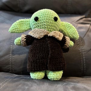 May include: A green crocheted plush toy with black eyes, wearing a brown coat with a tan collar. The toy is sitting on a gray couch.