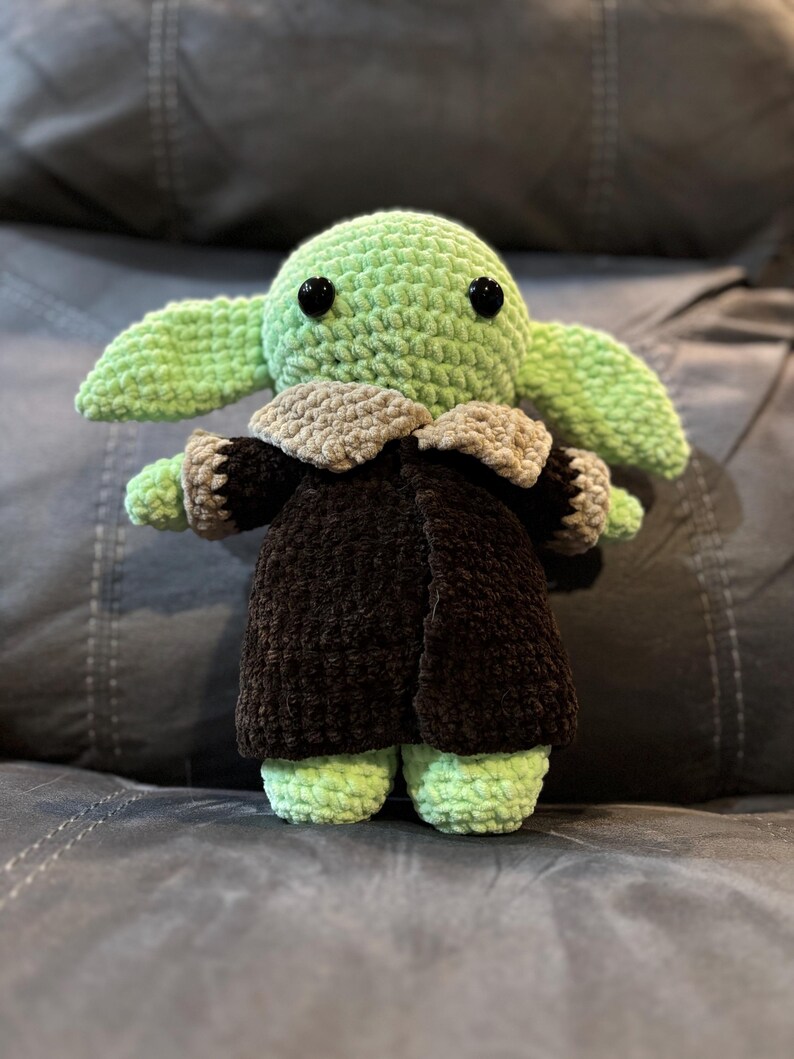 May include: A green crocheted plush toy with black eyes, wearing a brown coat with a tan collar. The toy is sitting on a gray couch.