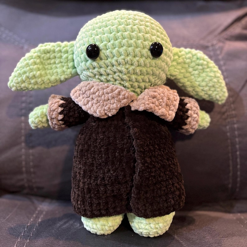 May include: A green crocheted plush toy with large ears, brown eyes, and a brown crocheted coat. The toy is wearing a brown crocheted coat with a tan collar.
