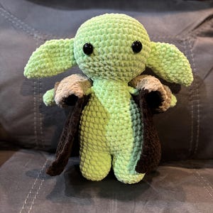 May include: A green crocheted plush toy with large ears and black eyes. The toy is wearing a brown and tan crocheted cape.