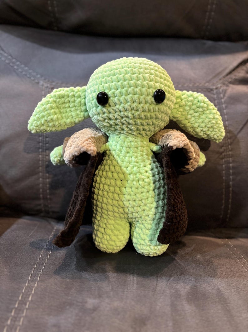 May include: A green crocheted plush toy with large ears and black eyes. The toy is wearing a brown and tan crocheted cape.