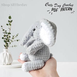May include: A gray crocheted elephant plush toy with white ears. The toy is sitting on a hand. The text "Cute Dog Crochet - PDF PATTERN" is visible in the image.