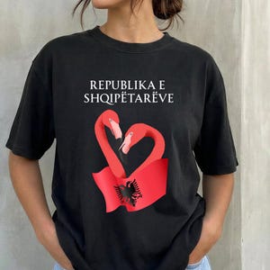 May include: A black short-sleeved t-shirt featuring the white text REPUBLIKA E SHQIPËTARËVE above a graphic of two pink flamingos forming a heart shape over a red flag with a black double-headed eagle emblem centred on the fabric.