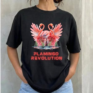 May include: A black short-sleeved t-shirt featuring a graphic print of two pink flamingos with spread wings standing in water. Below the birds, the text FLAMINGO REVOLUTION is printed in a red, slightly distorted, digital-style font.