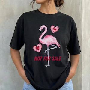 May include: A black short-sleeved t-shirt featuring a pink flamingo graphic centred on the chest. Two pink hearts are positioned near the flamingo, each containing a black double-headed eagle emblem. The text NOT FOR SALE is printed in red.