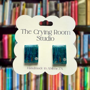 May include: A pair of earrings featuring miniature book covers of "Madeline" with a blue and green Eiffel Tower design. The earrings are on a white card with the text "The Crying Room Studio" and "Handmade in Abilene, TX".